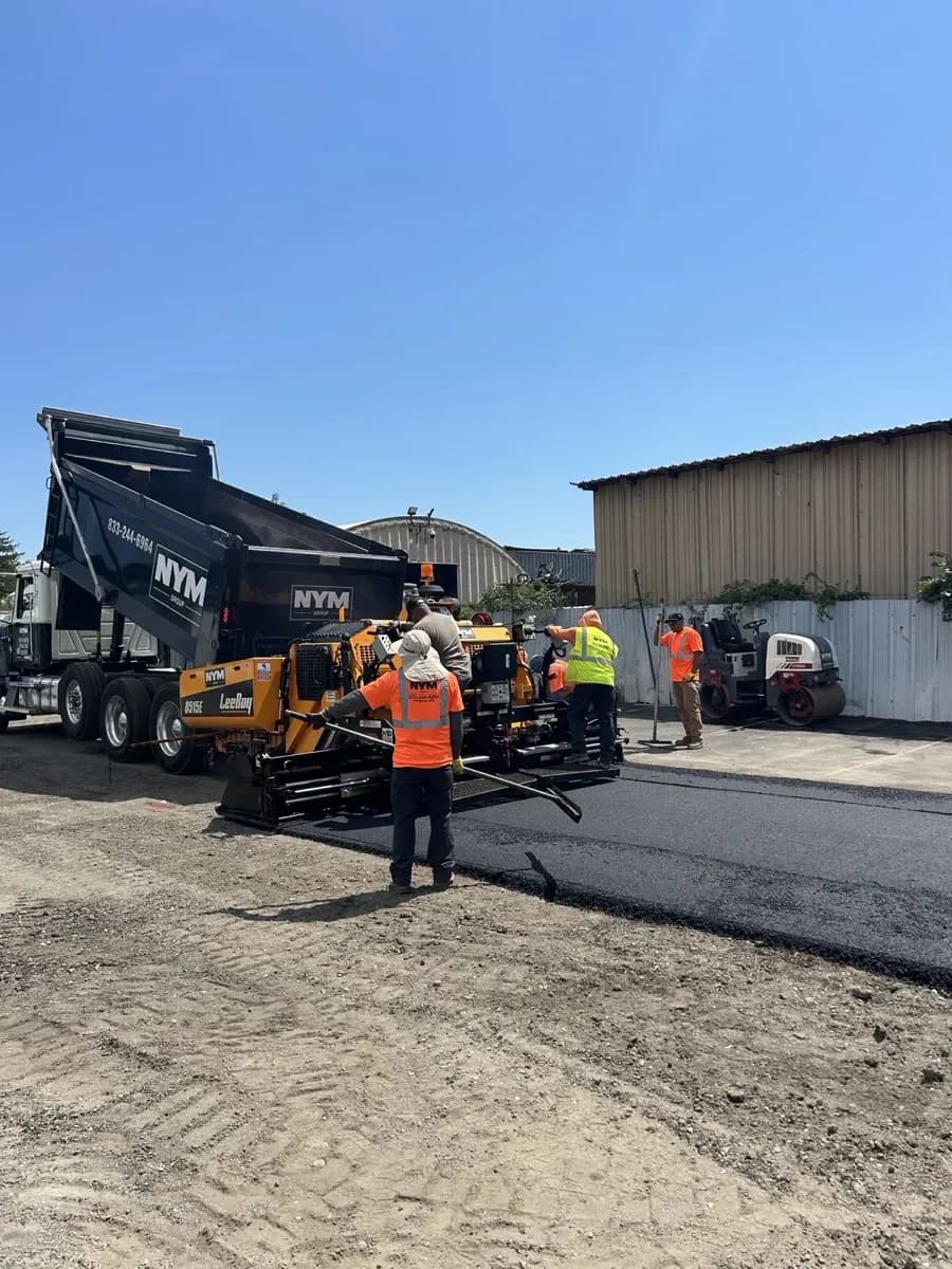 NYM crew in company hi-vis laying fresh asphalt with an NYM dump truck and paver